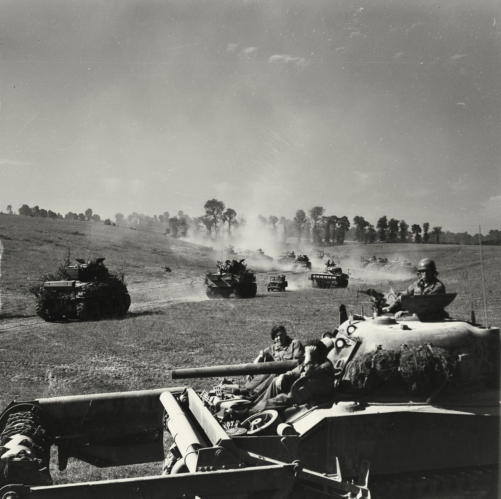 Black-and-white photograph of Allied tanks driving through rolling landscape with line of trees in distance. The tank in the foreground has equipment for a minesweeping flail in the front, with a helmeted soldier in the turret and two seated soldiers on either side of the gun. 
