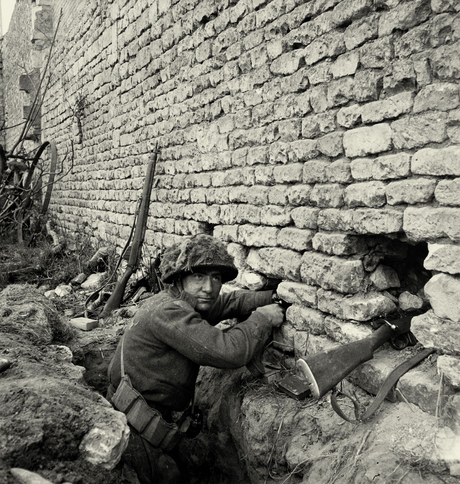 Black-and-white photograph of a helmeted soldier in a shallow trench, facing the camera while reaching into a high stone wall. The rifle beside him points through a narrow opening. In the background, a second rifle leans against the wall, with a metal wheel in the distance. 