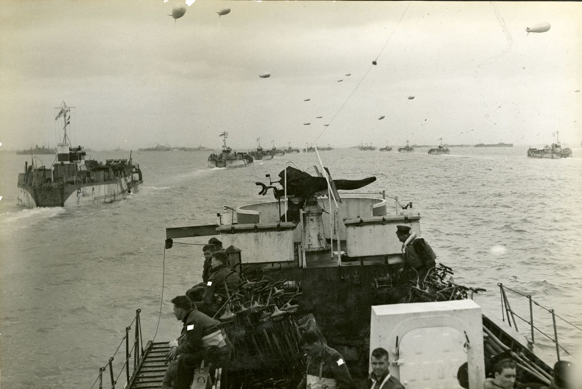 Black-and-white photograph of an overcast sky filled with barrage balloons. Two rows of landing craft head across the sea towards the horizon. On the landing craft in foreground, some soldiers look out over the rail at left, while others look up at the camera. 