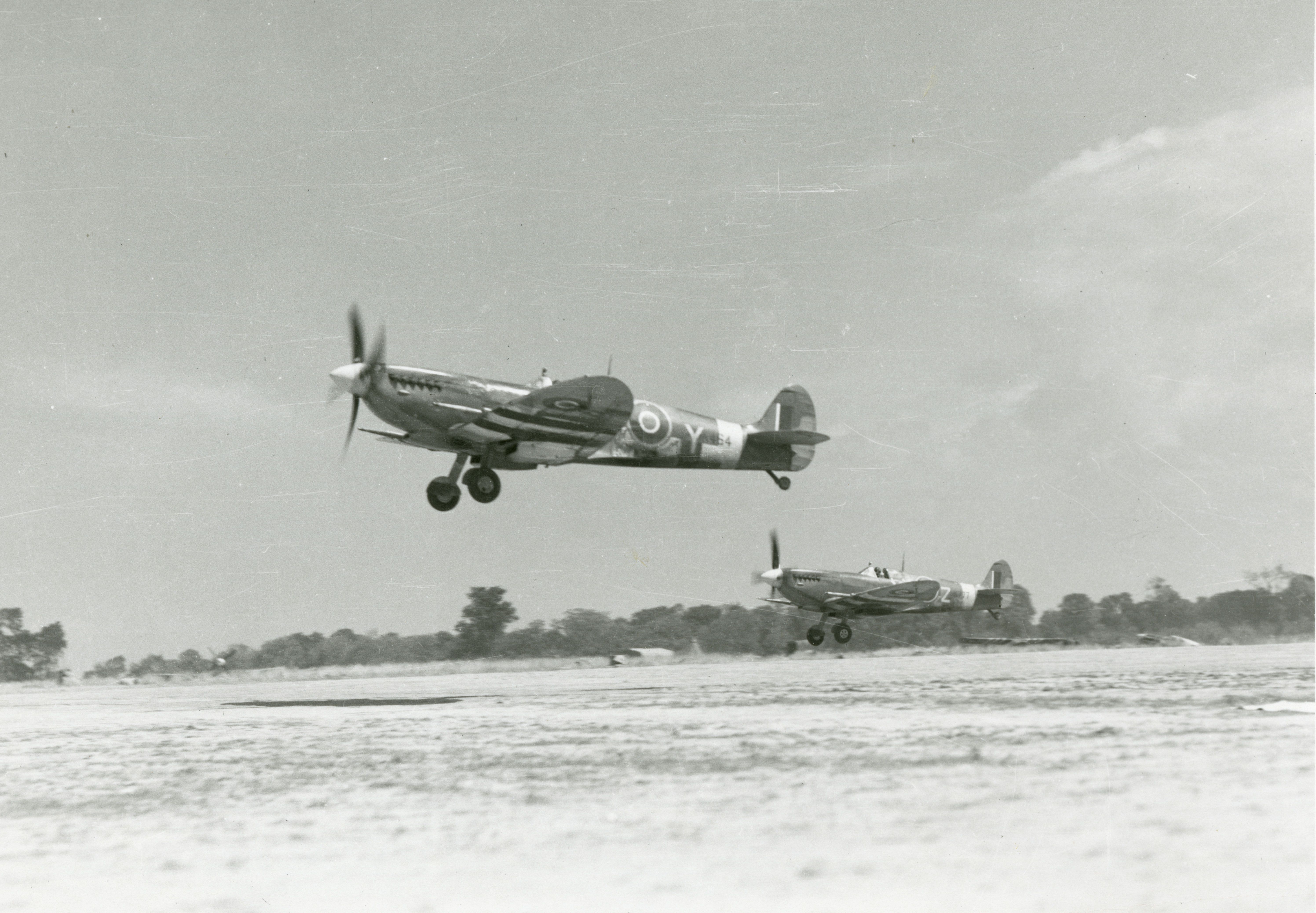 Black-and-white photograph of two Supermarine Spitfire fighter planes taking off from right to left over dusty open ground on a sunny day, with a line of trees in distance.