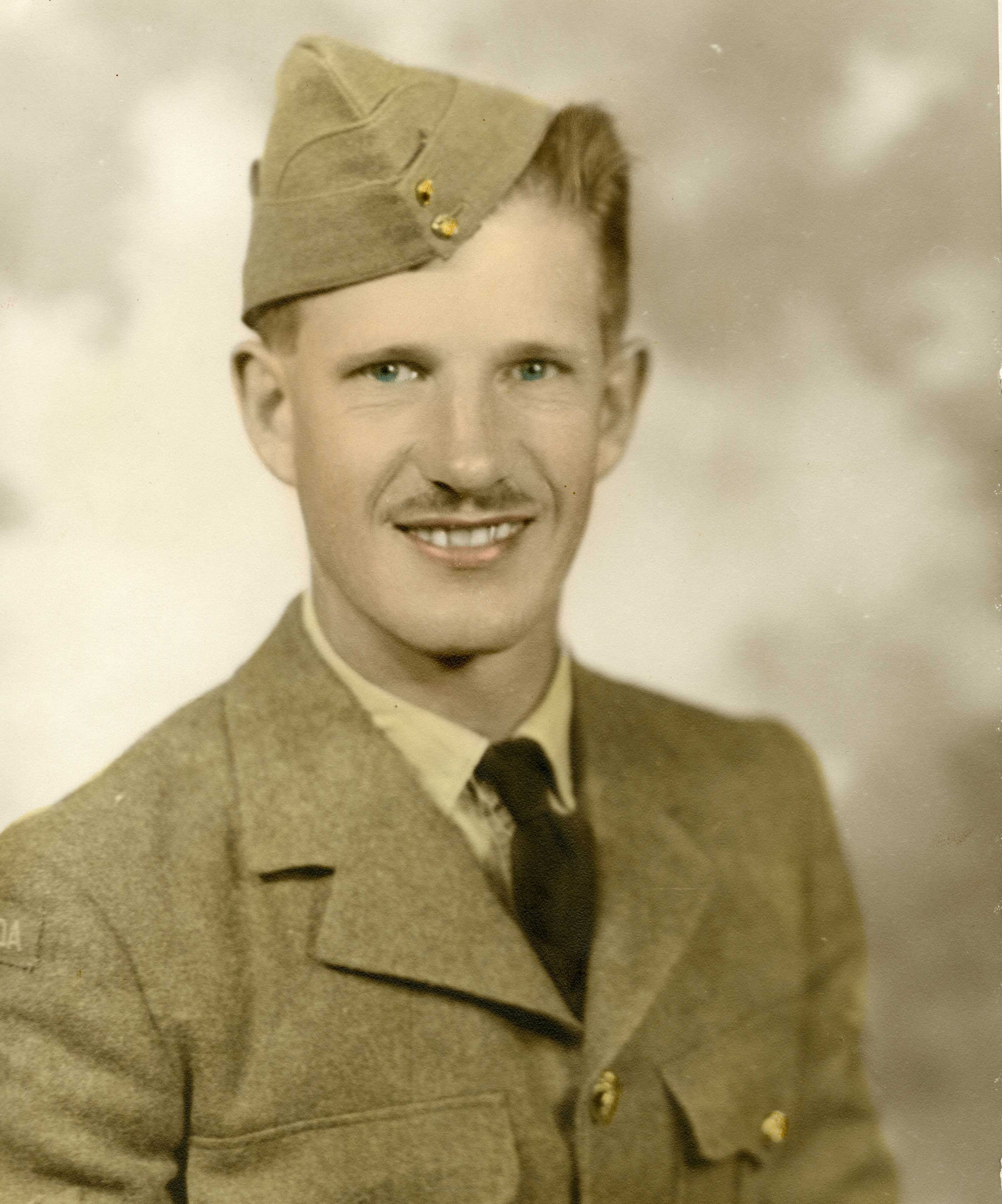 Hand-coloured black-and-white studio photograph of smiling man with a moustache in military uniform, wearing a necktie and a wedge-shaped cap perched at an angle. 