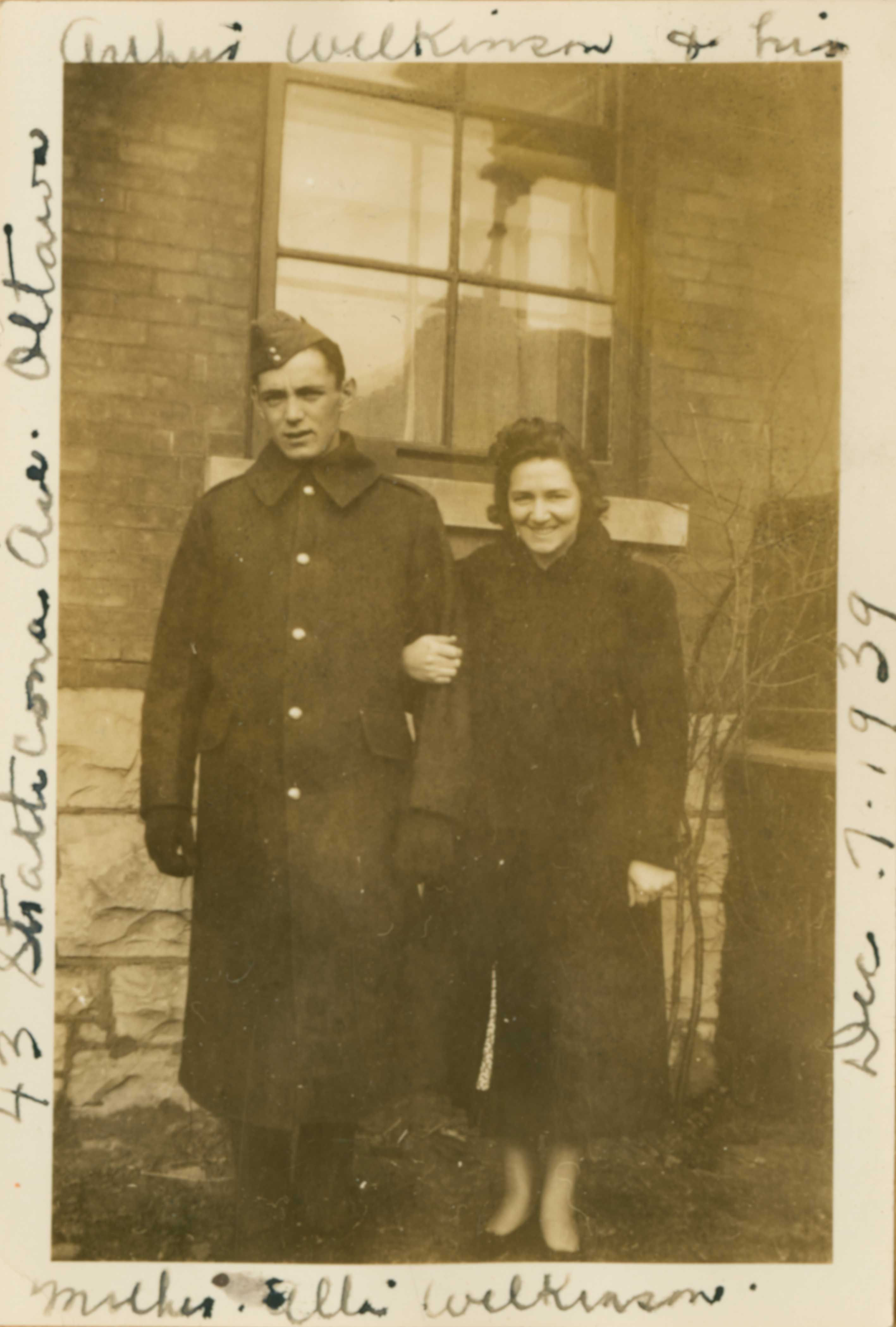 Sepia photograph outside a brick house of serious man on the left in military cap and greatcoat, standing beside a smiling woman on the right in dark coat with an arm tucked through his. Handwriting around the edges lists names, the place, and the date.
