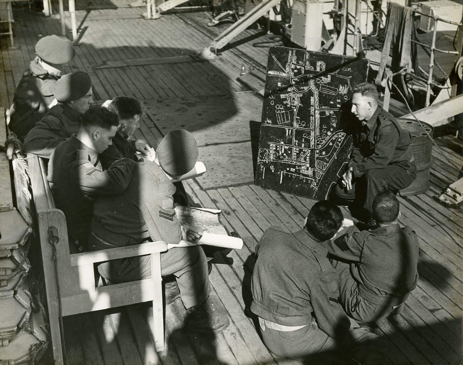 Black-and-white photograph of soldiers and officers in uniform at left and lower right, sitting on the deck and a wooden bench on a military ship, listening to a briefing by an officer crouched next to a blackboard at the upper right with a white chalk diagram.