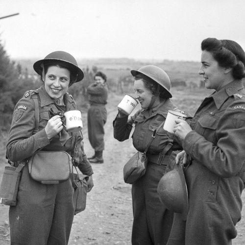 Three female Canadian army nurses in combat uniforms and Mark II helmets drinking tea outdoors.
