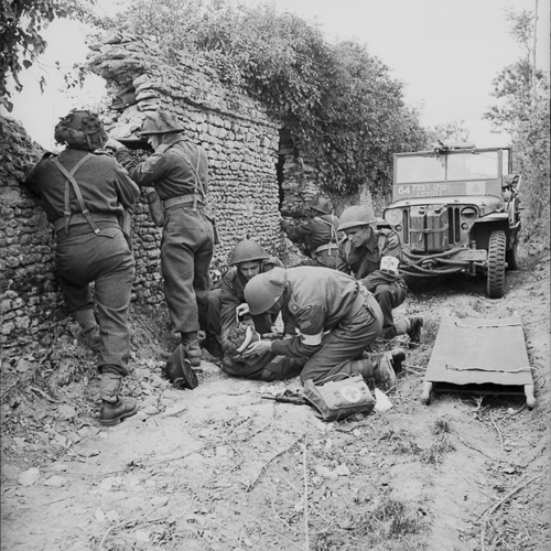 Three soldiers administer first aid to a wounded soldier. Three other soldiers defend their position from behind a rock wall.