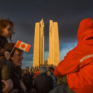 Two children sit atop the shoulders of adults as the Vimy monument stands, lit by the sun, in the background