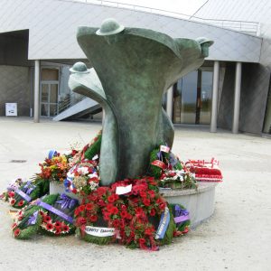 A stylized memorial statue with wreaths, flowers and poppies laid at its base..