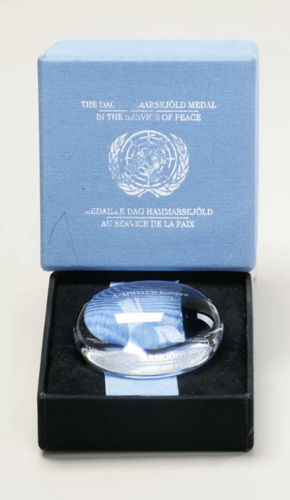 A clear crystal glass object in for the shape of a gently flattened sphere, with engraving, in a display box with the words 'The Dag Hammarskjöld Medal/In the Service of Peace/Médaille Dag Hammarskjöld/Au service de la paix' printed on the box lid.