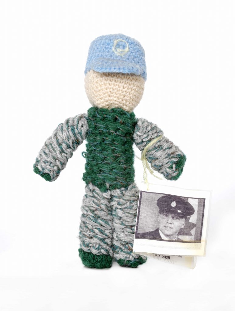 A knit doll wearing a light blue cap and green camouflage-style clothing. Attached to its arm is a tag showing the face of a uniformed man.