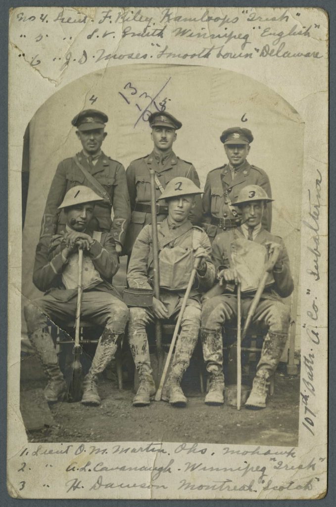 Three uniformed men stand behind three seated men wearing mud-stained uniforms and holding shovels and walking sticks. The men have been identified with numbers and accompanying text.