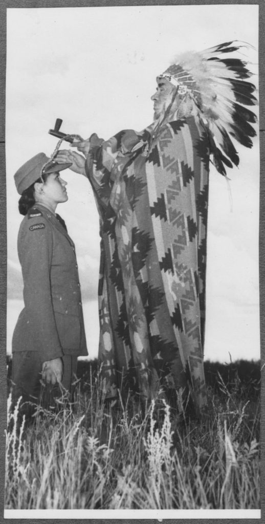 A uniformed Indigenous woman kneels in a field before an Indigenous man with mismatched ceremonial clothing and items.