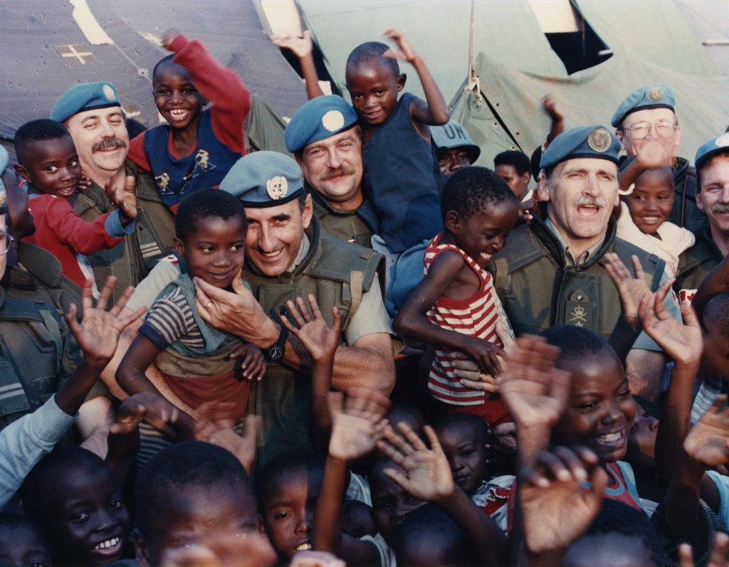 A crowd of smiling Black children and white men wearing Peacekeeping uniforms smile and wave for the camera