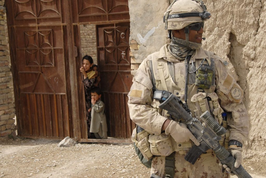 A uniformed man holding a rifle surveys his surroundings, while behind him, children peek out of a large wooden door.