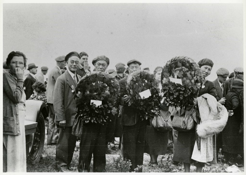 Three Japanese Canadian men and one Japanese Canadian woman hold wreaths and look to the camera while standing in a crowd