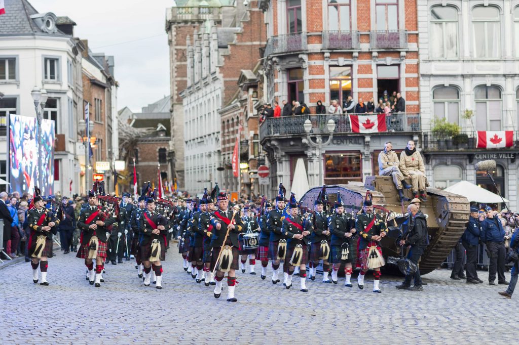 Men wearing ceremonial uniforms march down a street in Mons, alongside buildings decorated with Canadian flags. Two men sit atop of First World War-era tank.