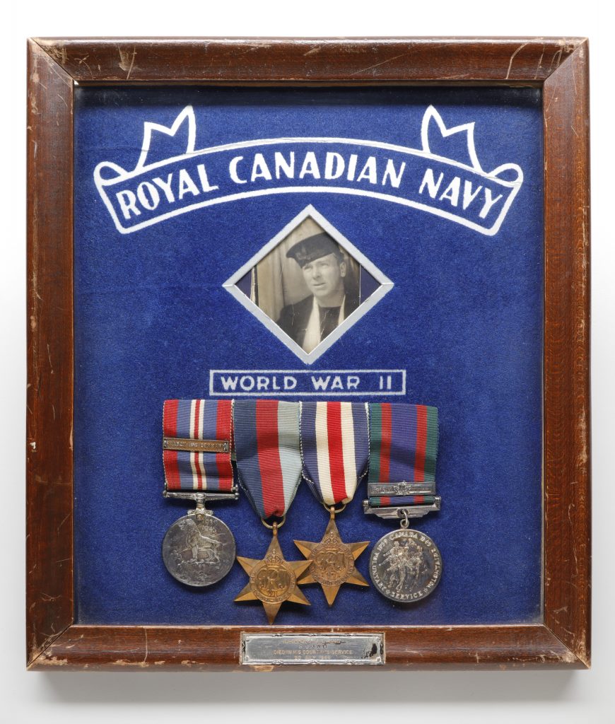 A black-and-white photograph of John Laing in uniform with his medals displayed underneath. The objects are encased in a dark wood frame and set off against a blue background. The words, “Royal Canadian Navy” and “World War II” appear at the top and centre, respectively, within the frame.