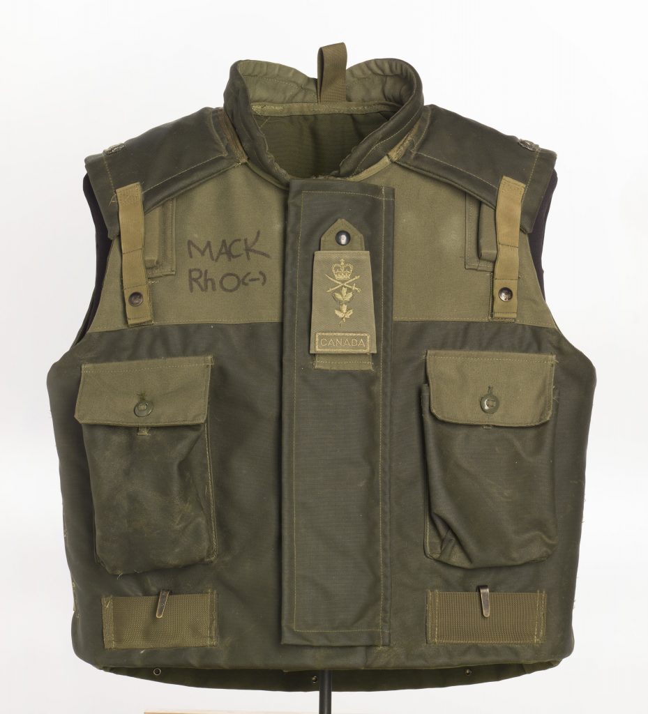 A green fragmentation vest with two large pockets and an embroidered badge at the centre of the chest featuring a crown, crossed swords, two maple leaves, and the word “Canada”.