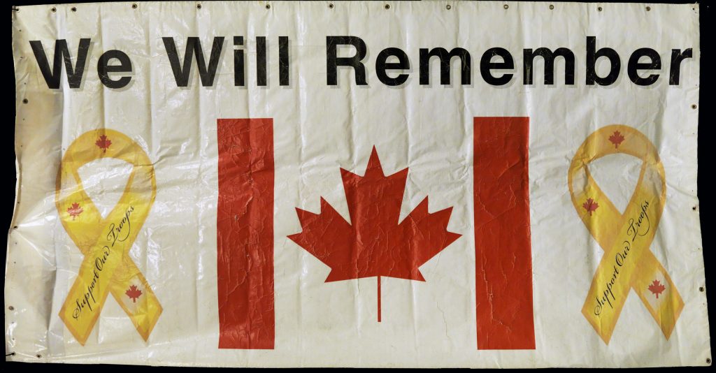 A worn white banner with metal grommets on three sides. The centre of the banner features a Canadian flag with yellow ribbons bearing the words “Support Our Troops” on each side. The words “We Will Remember” are displayed in large font across the top of the banner.
