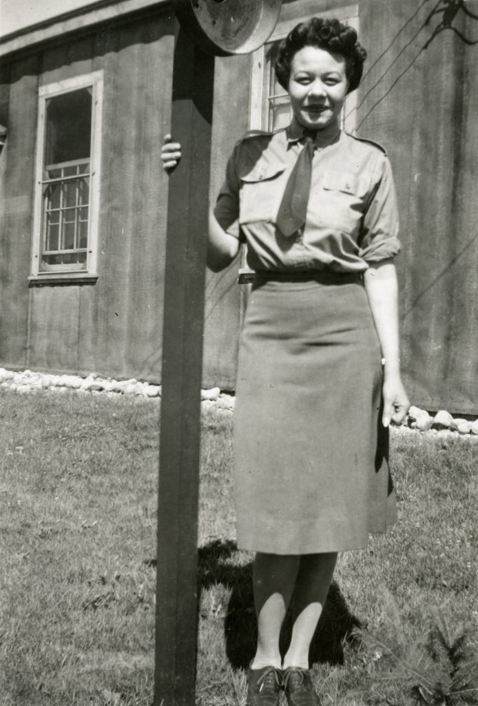 Minnie Gray in uniform, standing next to a wooden post, with a wooden hut and lawn visible in the background.