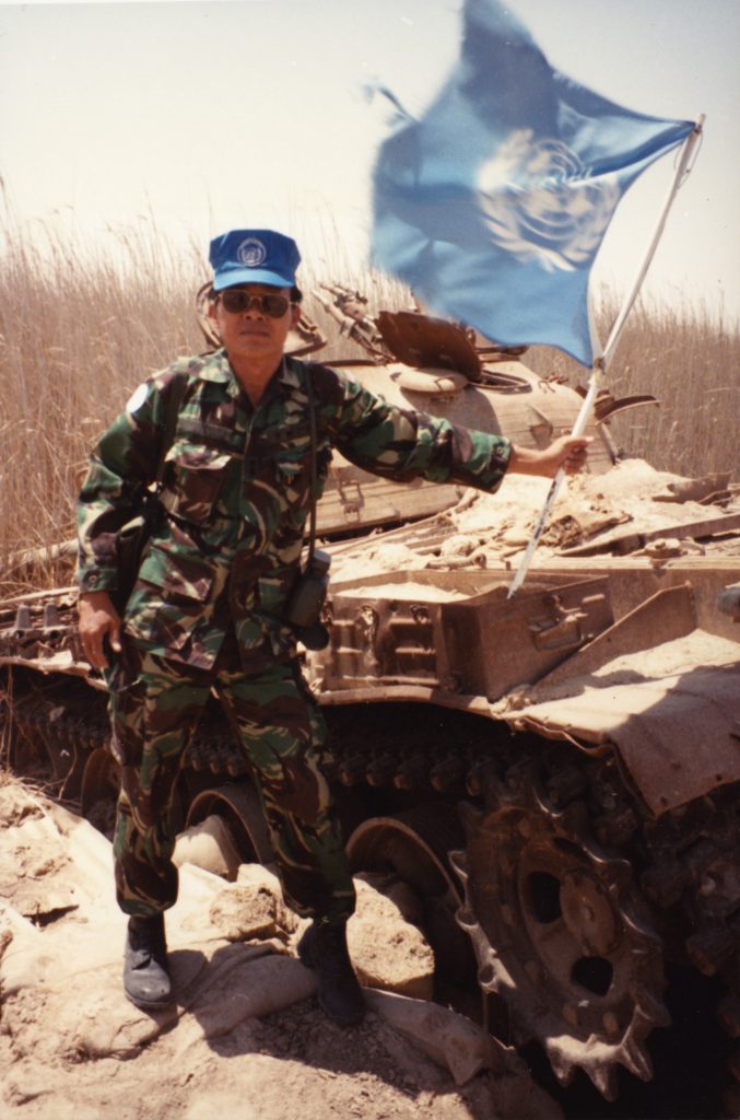 Ted Itani wears a camouflage uniform, a blue cap, and sunglasses as he hoists a blue and white United Nations flag. In the background, there is a destroyed tank.