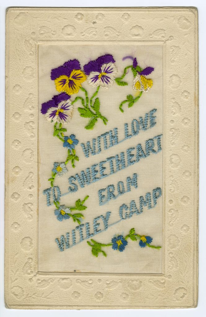 A cream-coloured card with an embossed border. The words “With love to sweetheart from Witley Camp” are embroidered in light blue thread and are accompanied by blue, violet and yellow flowers with green vines.