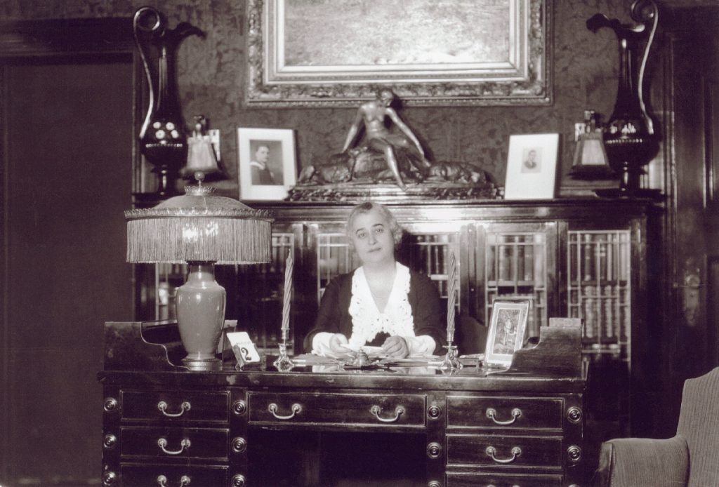 Lillian Bilsky Freiman sitting at her desk, surrounded by a large lamp, several picture frames, a statue of a reclining figure, and two large vases.