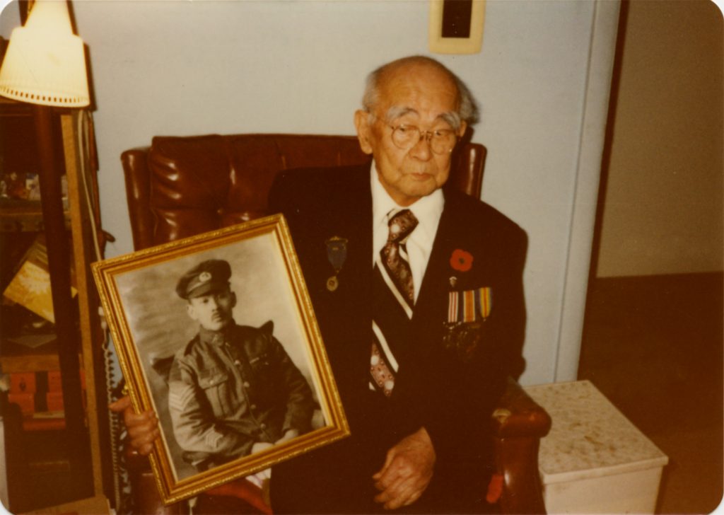 An elderly Masumi Mitsui wearing a Royal Canadian Legion uniform. He holds a black-and-white framed photograph of himself as a young soldier.