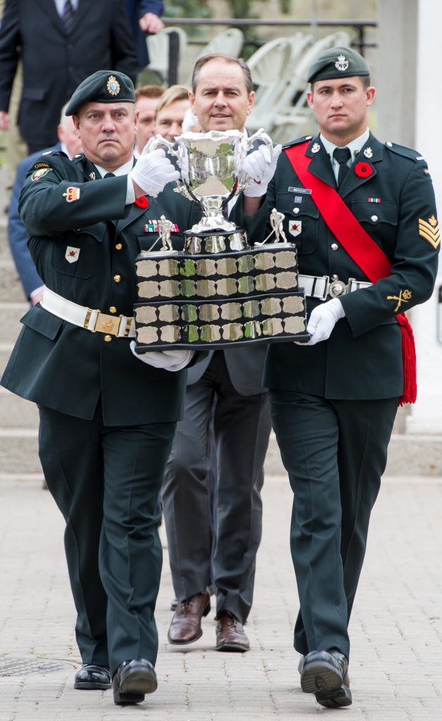 A procession led by two uniformed soldiers wearing a poppy on their lapel and carrying the Memorial Cup.
