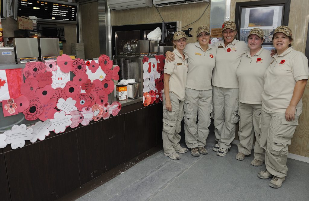 Five workers dressed in Tim Hortons uniforms and wearing poppies stand smiling with their arms around each other in a room decorated with red paper poppies and letters.