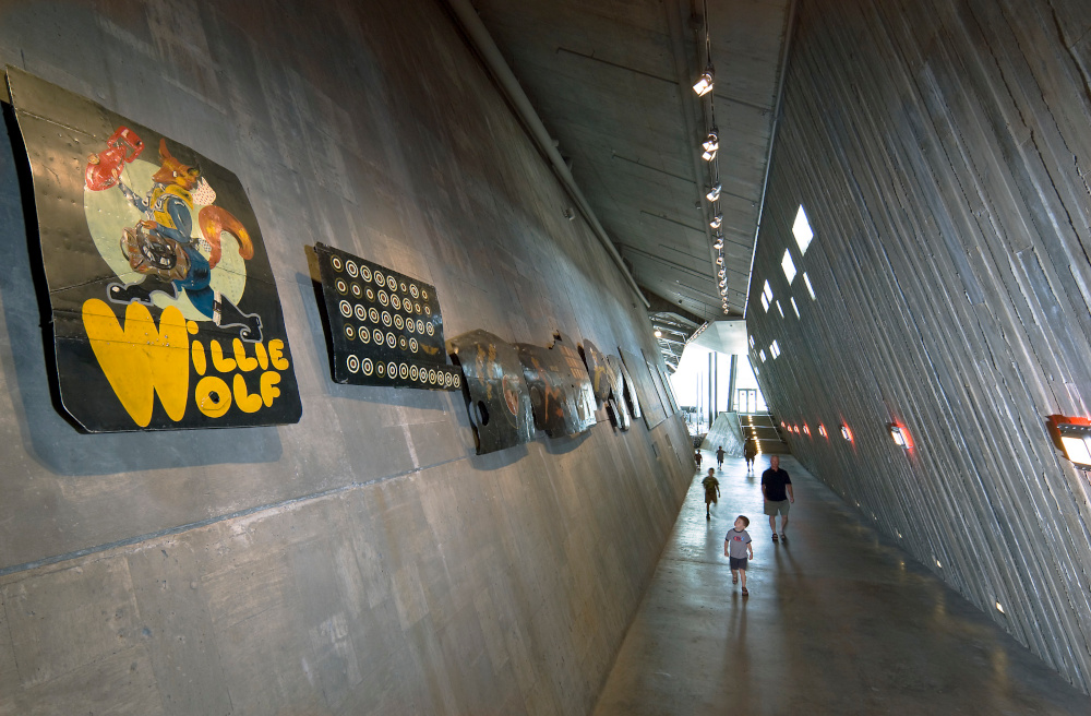 A long, tall concrete hallway with steeply slanted walls and an angled roof leads to a large sunlit room in the distance. There are paintings hung high up on the left wall.