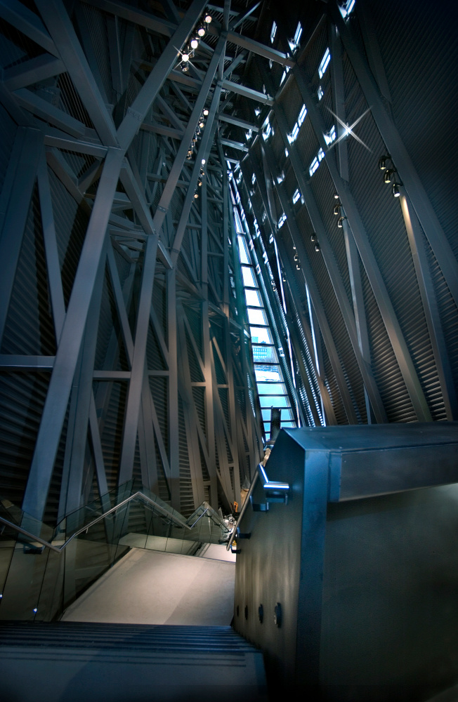 A very tall room with a visible structure of large metal posts and beams, a floor-to-ceiling narrow triangular window on the far wall, and descending concrete stairs in the foreground.
