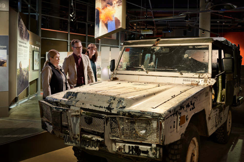 Three people stand in a museum gallery, looking at a battered white Jeep with bullet holes in its windshield.