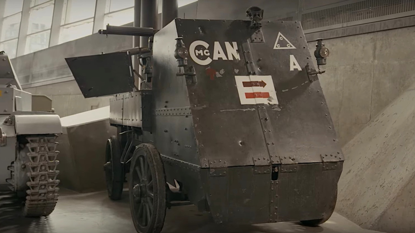 A dark green vehicle enclosed by tall, flat, metal plating, with two machine guns on the top and white markings painted on the front.