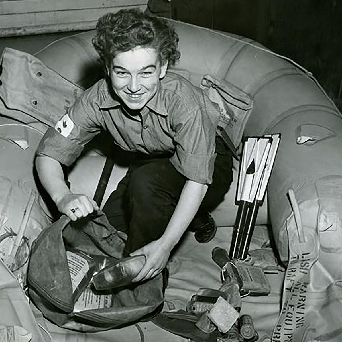 Photograph of a person kneeling in an inflatable raft, packing supplies into a bag. She is looking up and smiling at the camera.