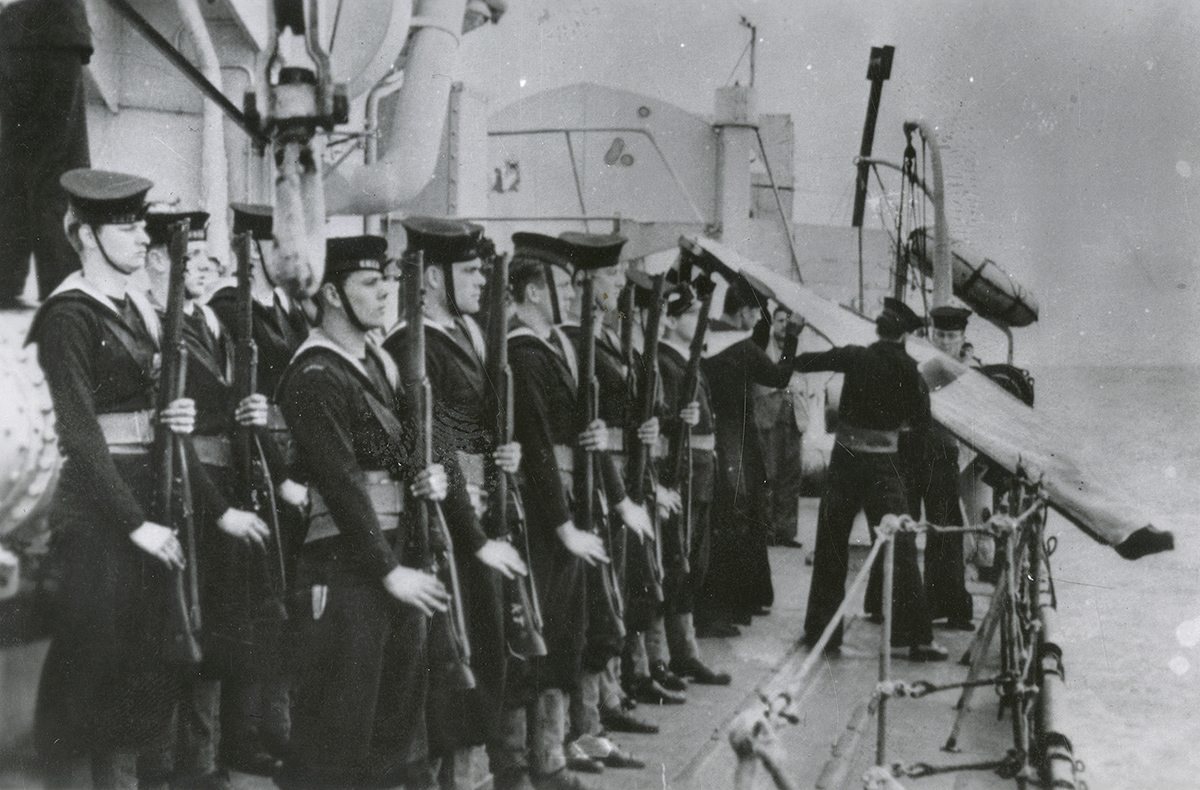 Photograph of sailors holding rifles and standing in formation on a ship deck. Two sailors are tipping a body in a shroud over the side of the ship.