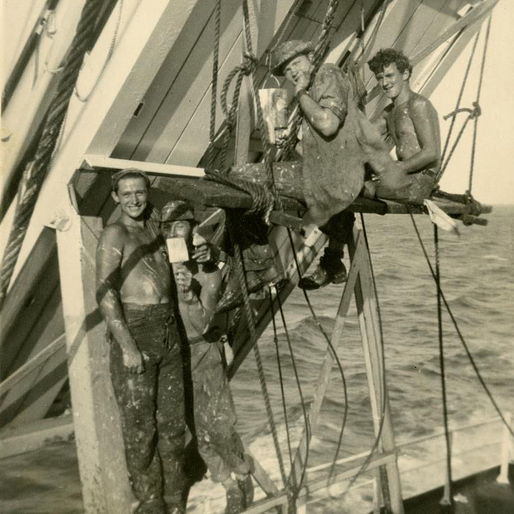 Photograph of four smiling sailors, covered in paint splashes while painting a life raft.