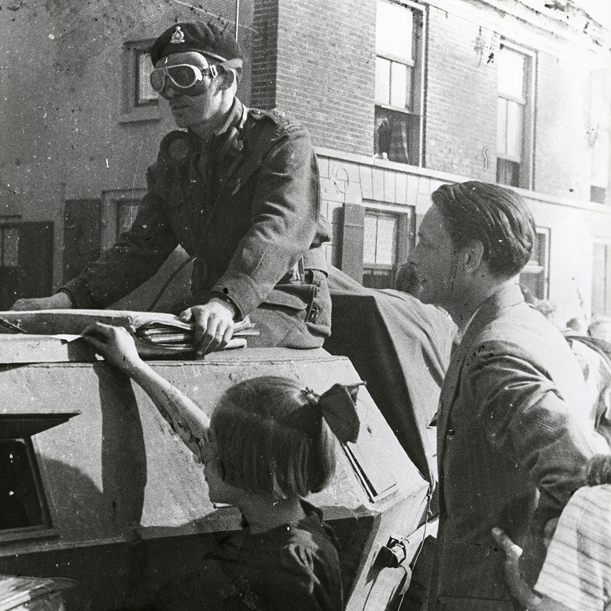 Photograph of an officer wearing goggles and a beret sitting atop an armoured car. An adult and a child stand to the side of the vehicle.