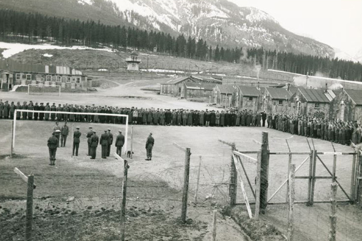 Photograph of an interment camp with prisoners standing around a field in the foreground, huts in middle ground and mountains in background.