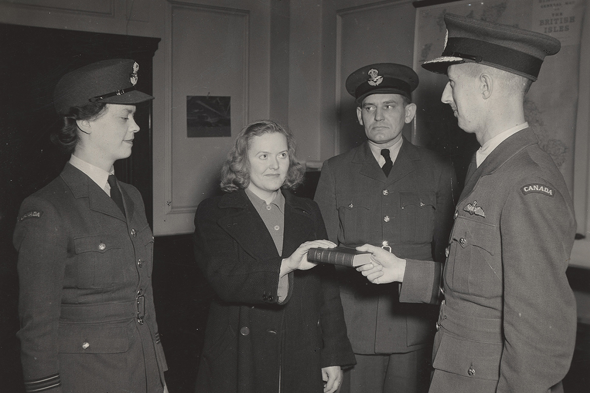 Photograph of a woman with her outstretched hand on a book, with two people in uniform standing beside her. The book is being held by a third person in uniform.