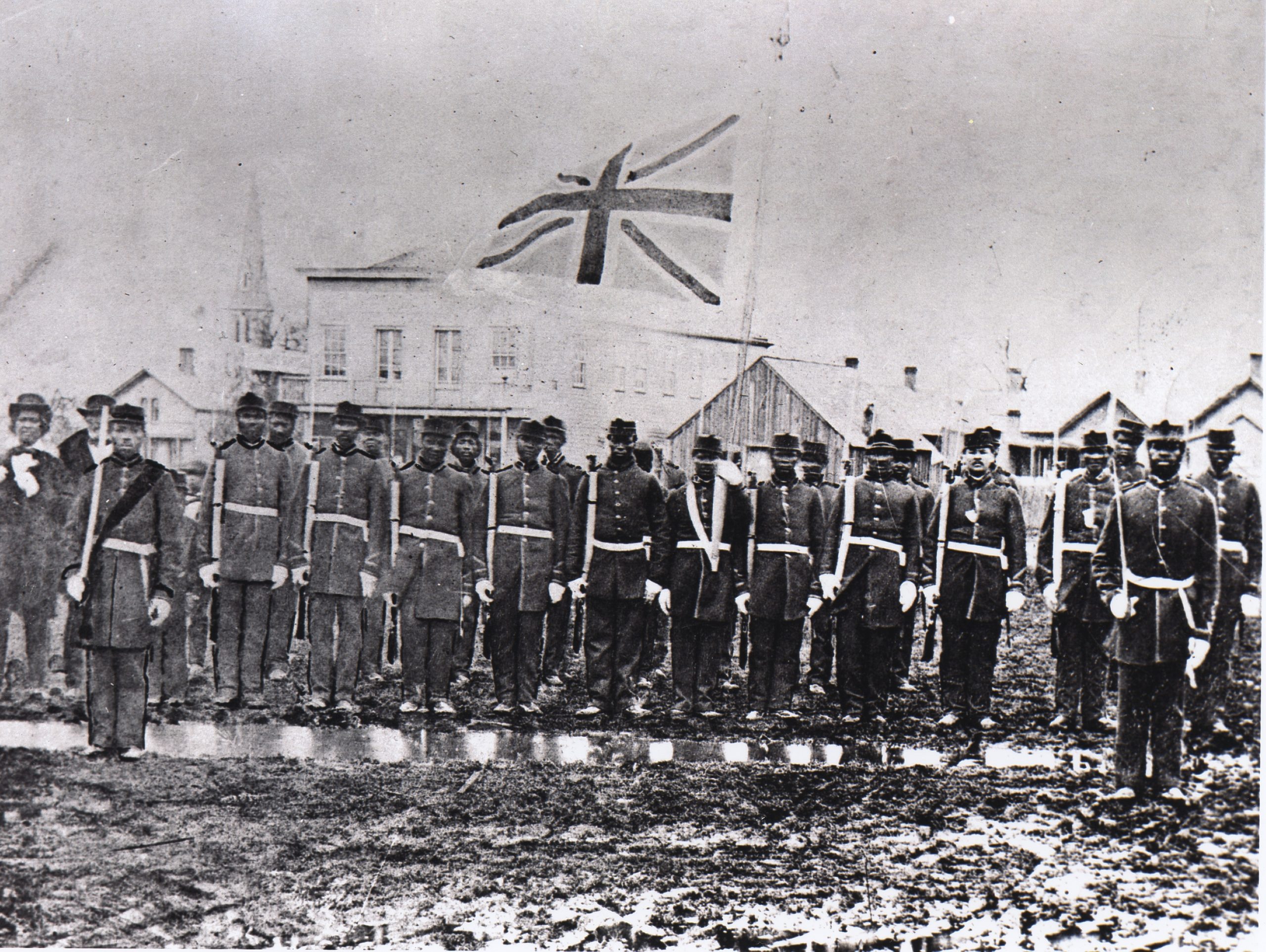 Black and white photo of black soldiers in a line. The Union Jack flag fliest above them.