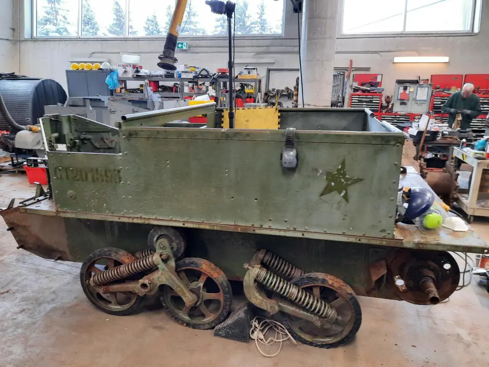 The same armored vehicle in a large garage filled with tools. Its tracks have been removed, darker paint shows where its markings have been removed, and its wheels are visibly rusted.