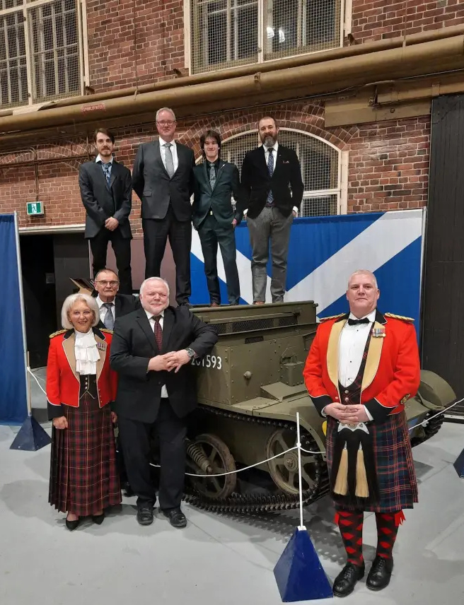 Several people in formal dress, including two in bright red jackets and tartan kilts, pose standing around and atop a low, square armoured vehicle.
