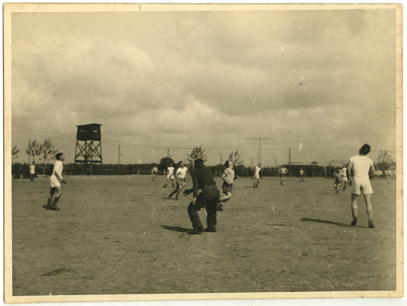 Black-and-white photo of several people, most in white shorts and t-shirts, spread out across an open field and looking upward towards a soccer ball high in the air. A dark, square guard tower and a line of fencing is visible in the background.