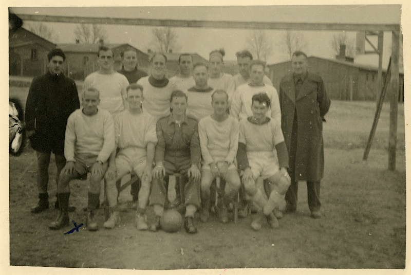 Black-and-white team photo of 14 light-skinned people with very short hair, standing and sitting. Most wear white shirts and shorts. Some are conspicuously thin and gaunt.