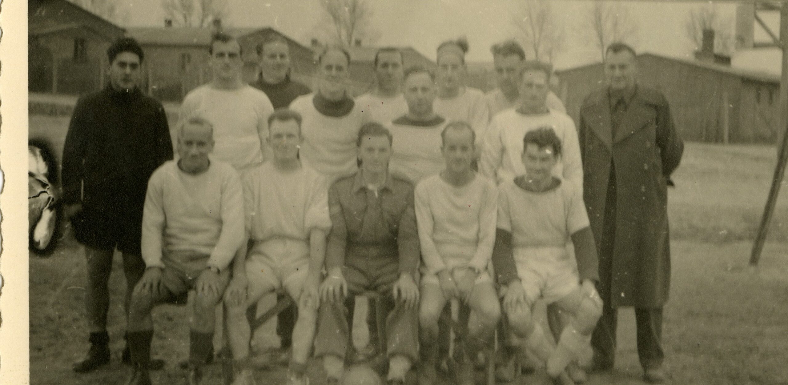 Black-and-white team photo of 14 light-skinned people with very short hair, standing and sitting. Most wear white shirts and shorts. Some are conspicuously thin and gaunt.