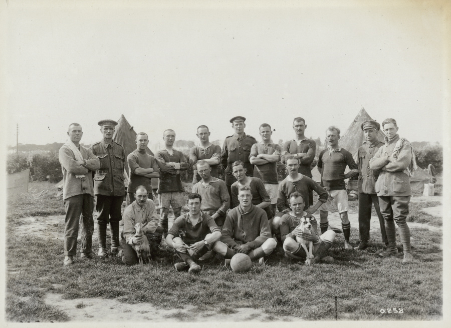 A group of 18 people pose in rows behind a soccer ball. Some wear shorts and short-sleeved shirts, others military uniforms. At the far left of the front row, a smiling man holds a young goat; to the far right, a man holds a small dog in his lap.