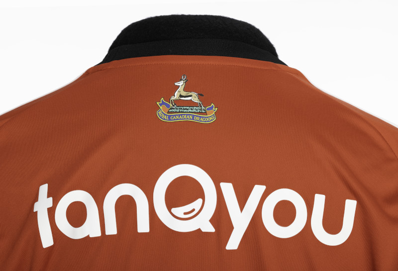 Close up on the mid-back of an orange-brown, short-sleeved athletic shirt with a logo of an antelope above a banner reading “Royal Canadian Dragoons” and large white text reading “tanQyou.”