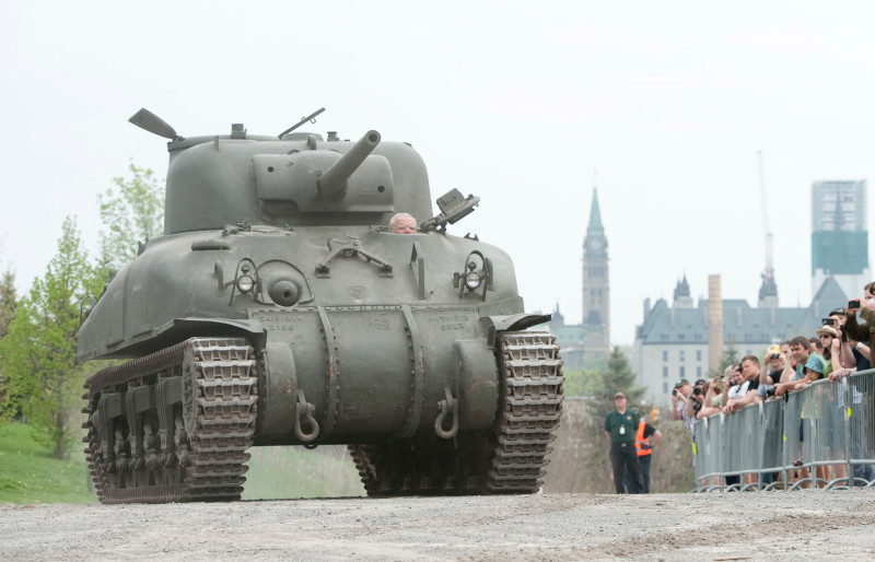 A large, green military tank driving past a crowd of people.