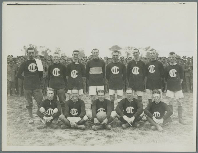 Black-and-white team photo of eight light-skinned people with very short hair standing behind five others who sit on the ground. The middle seated person holds a soccer ball. Most wear light shorts and dark shirts with a prominent logo. A large crowd of people in military uniform fills the background behind them.