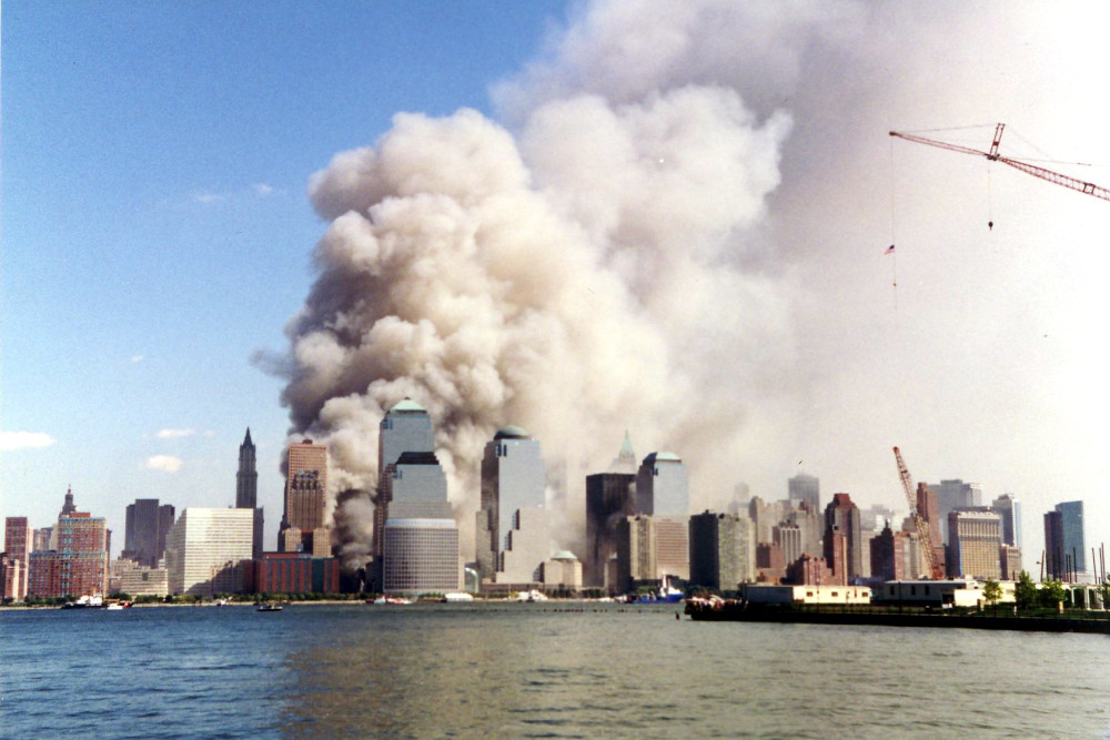 A huge cloud of smoke billowing up behind a city skyline with a body of water in the foreground.]